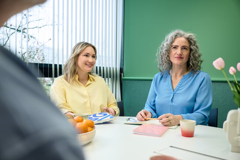 Two women are sitting at a table in a bright room, talking. One is wearing a yellow top and smiling, the other is wearing a blue top.