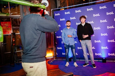 Twee mannen poseren voor een blauwe achtergrond met ‘bol’-logo's, terwijl een fotograaf hen vastlegt.