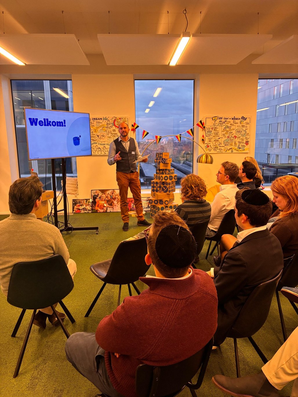 A man gives a presentation in a modern office with large windows. Participants sit on chairs and listen attentively.