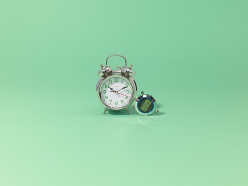 Two metal clocks, one large and one small, stand on a mint green background. The large clock shows 10:10, while the small one has a digital display.