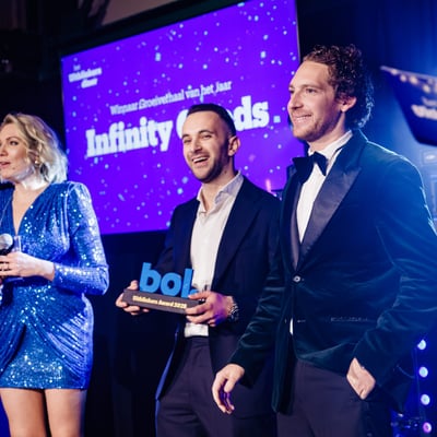 Three people in formal attire on stage. A woman in a blue sequined dress holds a microphone. Two men are smiling, one of them holding an award. Text is displayed on a screen in the background.