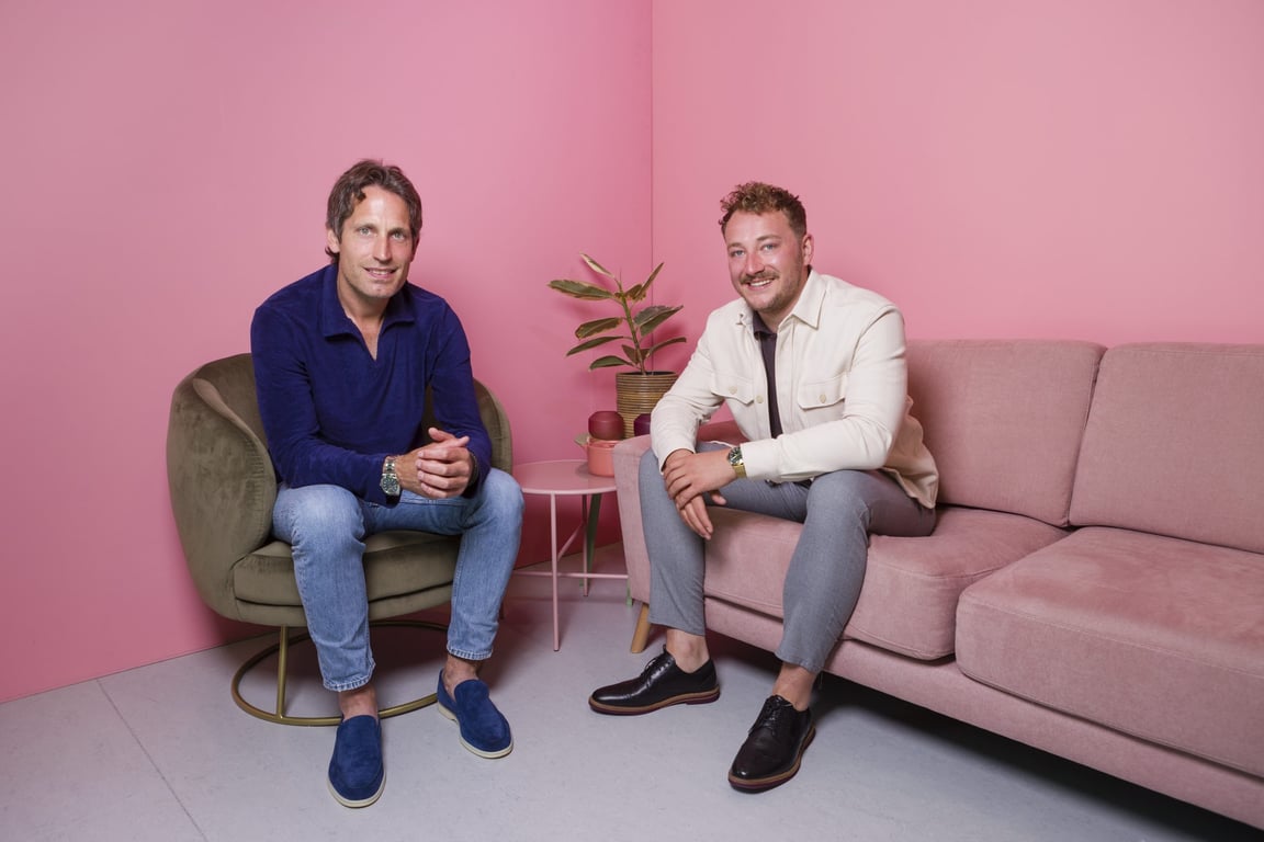 Jochem Hes en Jasper van Wijk zitten op een bank met een roze achtergrond.