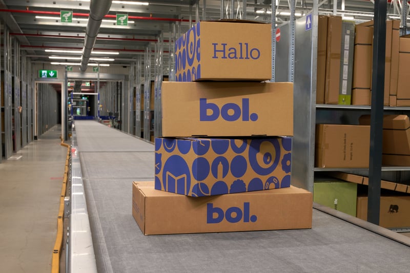 Een stapel van vier bol-pakketjes op een lopende band in een groot magazijn. De setting oogt industrieel en georganiseerd.