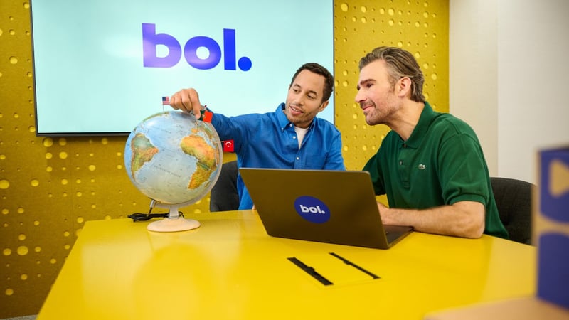 Twee vrouwen zitten aan een tafel in een lichte kamer met groene muren. De ene draagt geel, de andere blauw. Ze glimlachen en praten met elkaar.