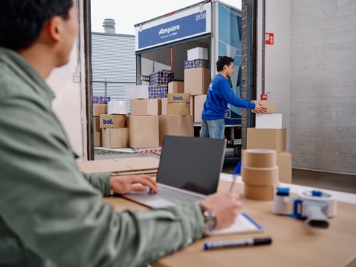 A man in a green coat sits at a table with a laptop and watches another man unload boxes from a truck in a warehouse setting.