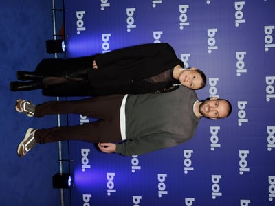 Een glimlachende man en vrouw poseren samen op een blauw tapijt voor een achtergrond met het merk ‘bol.’, wat een vrolijke en feestelijke sfeer uitstraalt.