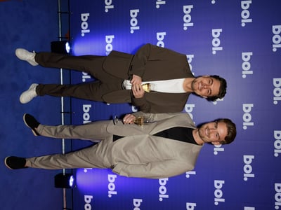 Two people in suits stand on a blue carpet, smiling and holding drinks. They pose in front of a backdrop with a repeated "bol." logo.