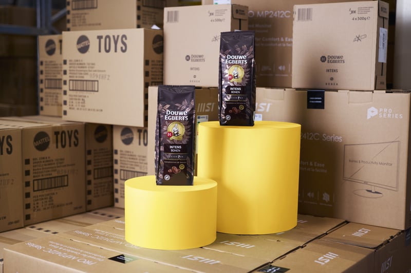 Two coffee bags stand on yellow cylindrical stands between stacked cardboard boxes on a wooden pallet, creating an industrial and vibrant presentation.