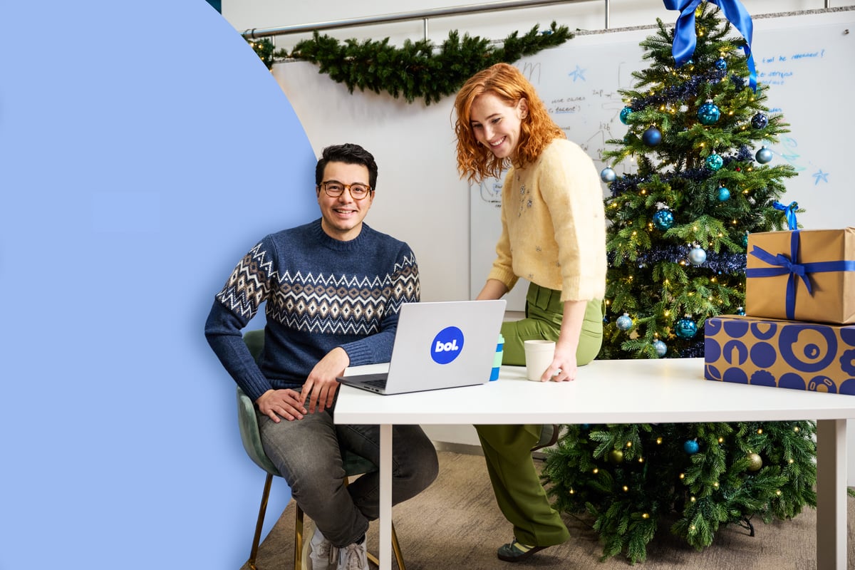 Een glimlachende man in een feestelijke trui en een vrouw in een licht vest zitten aan een tafel met een laptop, naast een kerstboom en cadeauverpakkingen, wat een vrolijke feeststemming creëert.