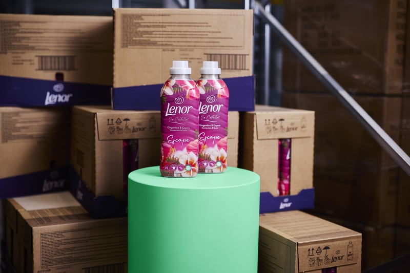 Twee flessen wasmiddel staan op een groene cilinder, omringd door gestapelde bruine dozen in een magazijnsetting.