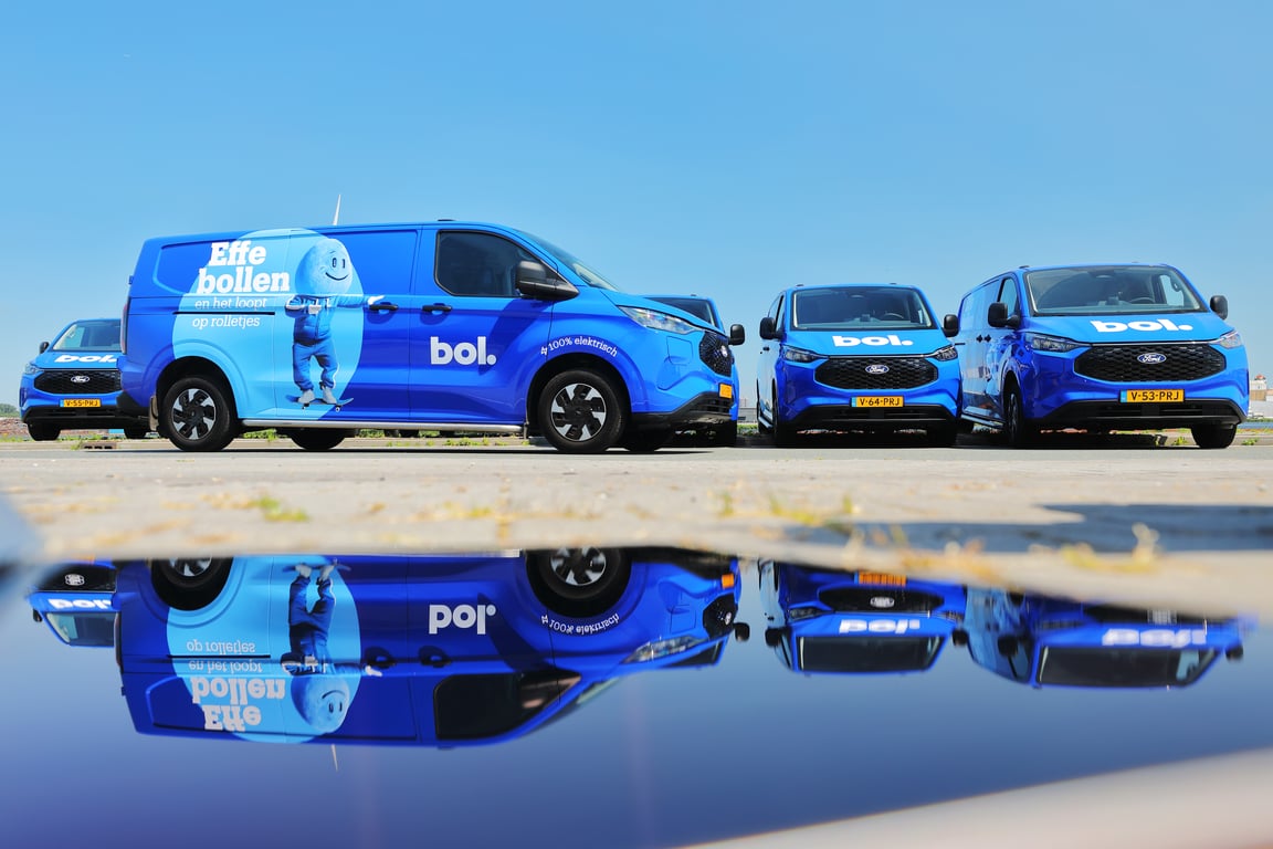Een blauw Ampère-busje geparkeerd langs de kant van de weg, omgeven door groen en woongebouwen.
