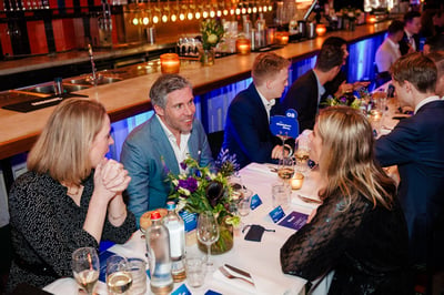 Mensen in formele kleding zitten aan een lange tafel versierd met bloemen en kaarsen in een levendige, warm verlichte restaurantsetting, waar ze geanimeerde gesprekken voeren.
