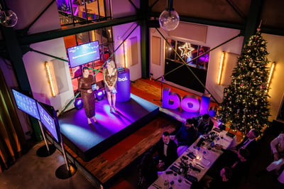 Een feestelijke scène waarin twee mensen op een met paarse lampen verlicht podium staan te praten tegen een publiek.