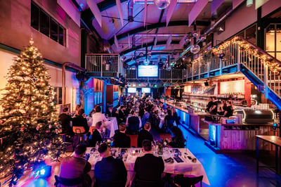 Feestelijk diner in een grote zaal met versierde kerstboom, elegante eettafels, warm verlichte bar en mensen die genieten van de levendige, feestelijke sfeer.