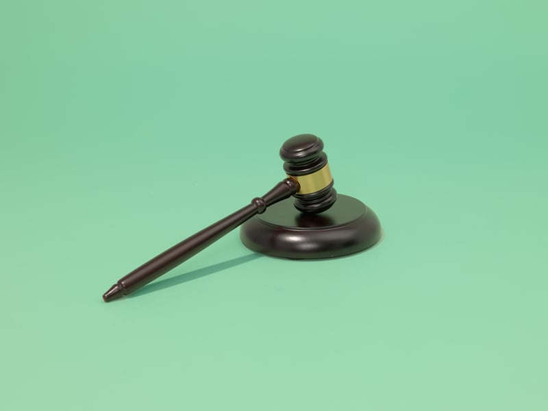 Een houten rechtershamer ligt schuin op het bijbehorende blok tegen een effen groene achtergrond, als symbool voor recht en gerechtigheid.