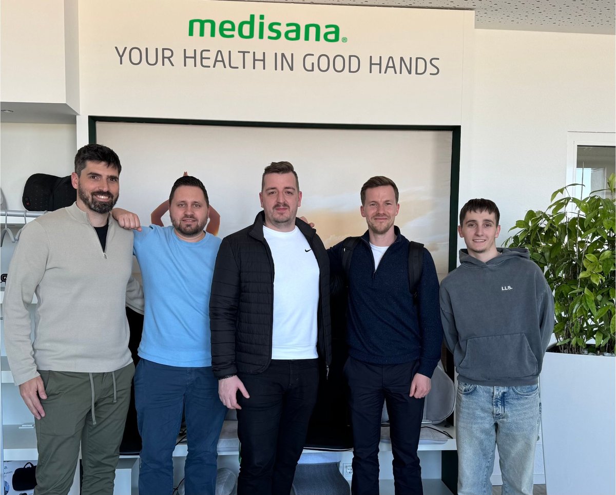 Five smiling employees stand next to each other under a sign that reads 'Medisana: Your Health in Good Hands'.