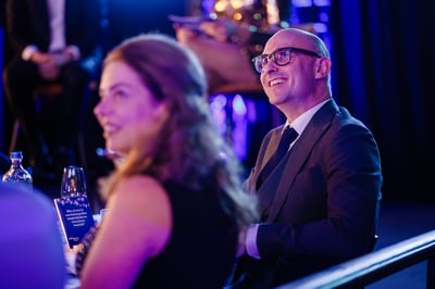 Een glimlachende man met een bril en een pak zit op een formeel evenement, zacht verlicht in blauwe en paarse tinten.