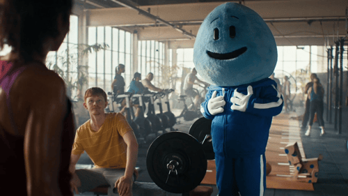 A person in a blue droplet mascot costume stands in a gym next to people using exercise equipment.