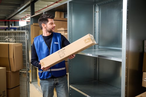 Een magazijnmedewerker in een blauw vest houdt een rechthoekig pakket vast. Hij kijkt naar lege metalen stellingen. Op de achtergrond staan kartonnen dozen. De sfeer is druk en gefocust.
