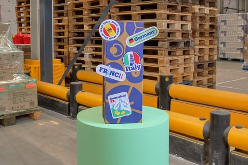 Een kartonnen doos met stickers van Europese landen, zoals Duitsland, Italië en Zwitserland, staat op een groene verhoging in een industrieel magazijn. Op de achtergrond zie je stapels houten pallets en veiligheidshekken.