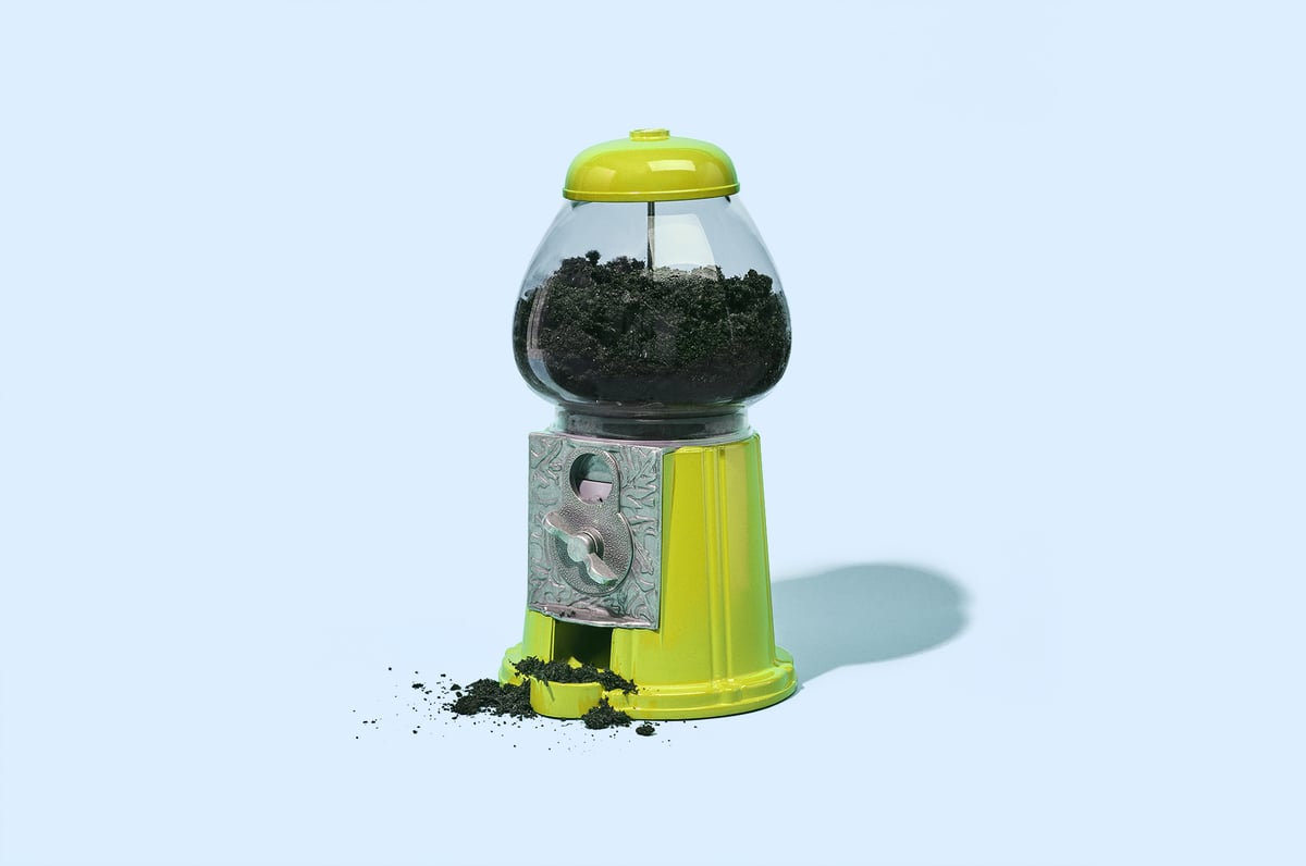 Een felgroene kauwgomballenautomaat gevuld met aarde, in plaats van kauwgomballen, staat op een lichtblauwe achtergrond.