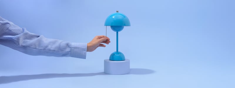 A hand pulls the chain of a small, modern blue lamp on a white base, against a blue background.