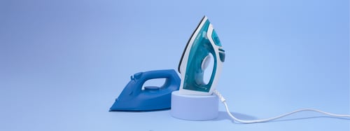 Een blauw strijkijzer en een blauwgroen en wit strijkijzer rechtop geplaatst op een lichtblauwe achtergrond. Het blauwgroene strijkijzer staat op een ronde voet, met een snoer dat naar buiten loopt.