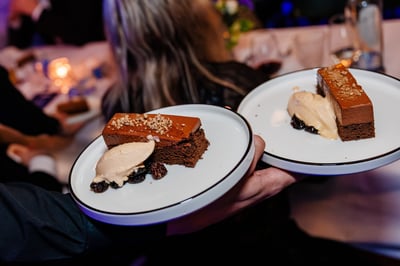 Er worden twee borden chocoladetaart met slagroom en bessen geserveerd. De setting is een elegant evenement met gedempte verlichting, wat een verfijnde sfeer uitstraalt.