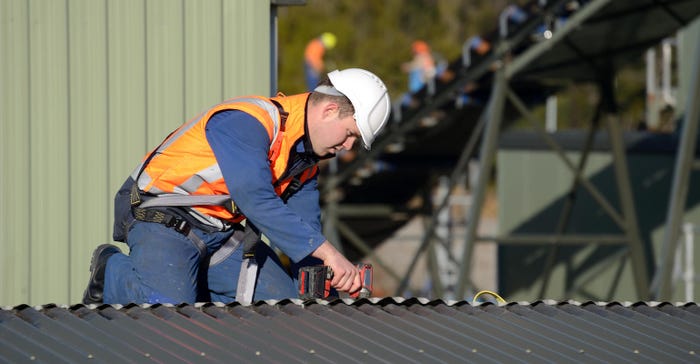 A builder wearing a safety harness for working at heights unscrews roofing iron on a building