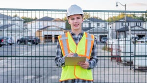 Young, smiling construction worker wearing safety gear and holding a clipboard