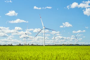 Image shows a wind power plant in the green field against cloudy sky.
