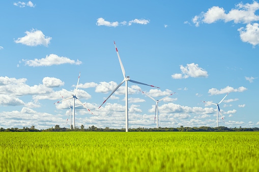 Image shows a wind power plant in the green field against cloudy sky.