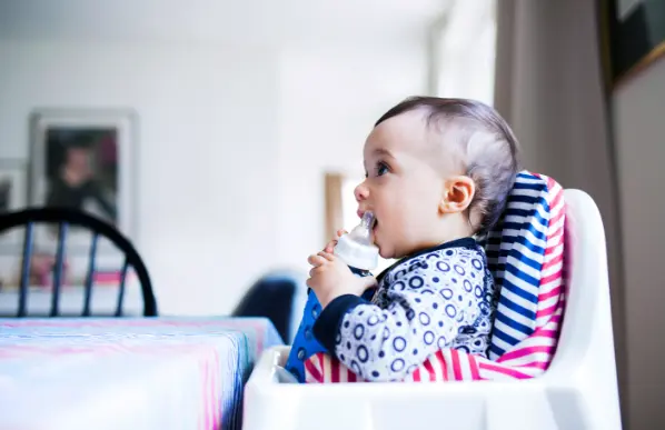 Baby in striped high chair eating with spoon, wearing patterned shirt, looking to the side in bright kitchen setting.