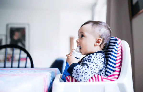 Baby in striped high chair eating with spoon, wearing patterned shirt, looking to the side in bright kitchen setting.