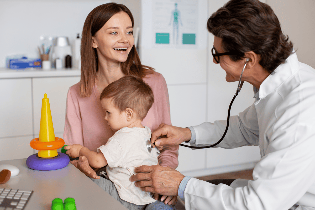 Doctor examining baby with stethoscope while smiling mother holds child in medical office with colorful toys nearby.