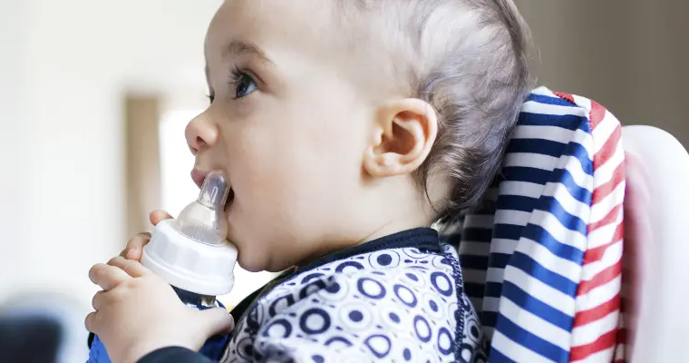 Baby in patterned outfit drinking from a bottle while sitting in a colorful striped high chair