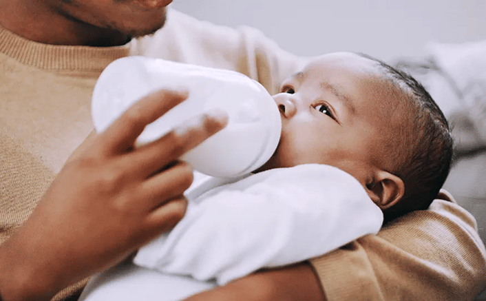 Father bottle-feeding an infant baby who is lying down and looking up while drinking from a white bottle