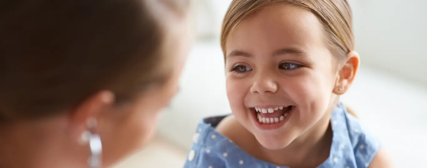 Young blonde girl with blue eyes smiling broadly while brushing her teeth, wearing a blue polka dot shirt in a bright bathroom.