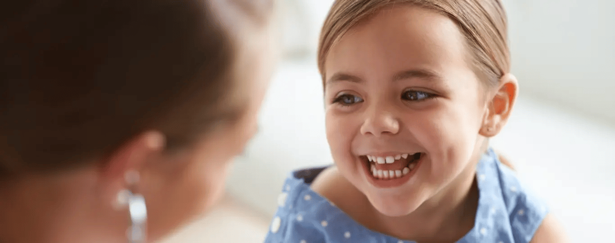 Young blonde girl with blue eyes smiling broadly while brushing her teeth, wearing a blue polka dot shirt in a bright bathroom.