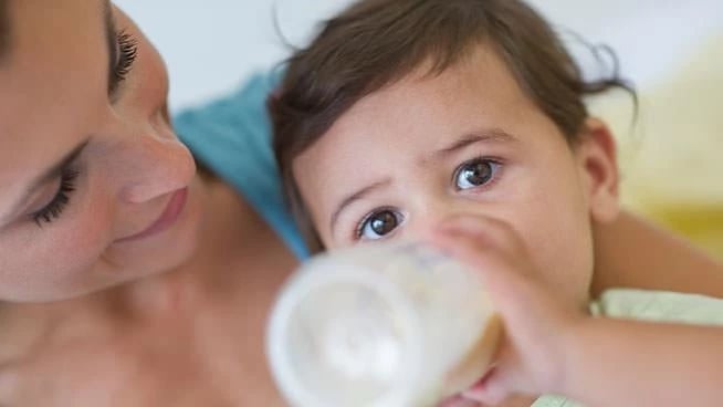 Mother holding baby who is drinking from a bottle, both looking at camera in bright indoor setting