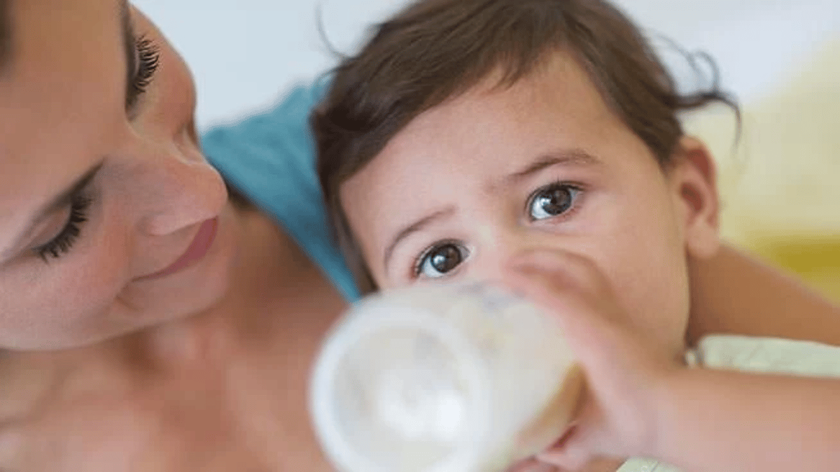 Mother holding baby who is drinking from a bottle, both looking at camera in bright indoor setting