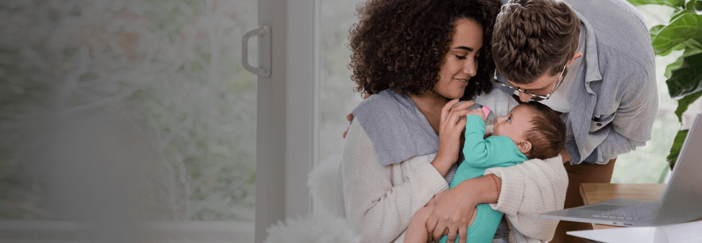 A couple with curly-haired woman and man in glasses lovingly look at their baby while sitting at a table with a laptop in a bright home office.