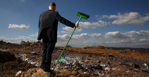 A man with a broom looks over a landfill, signifying a desire or need to clean it up.