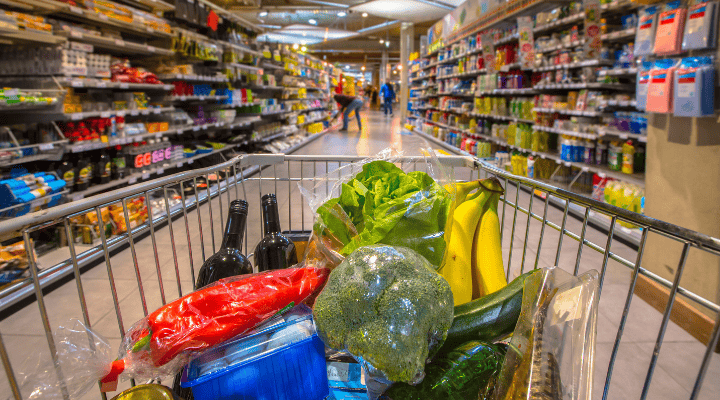 Een winkelwagen vol kleurrijke groenten staat in een goedgevulde supermarktgang.