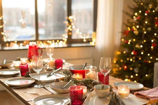 Mesa de Navidad con vasos rojos de cristal, copas y platos junto ventana con luces navideñas al fondo