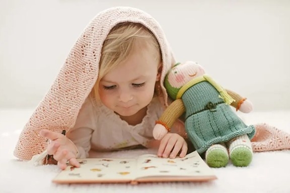 Child reading a book with a plush toy under a cozy blanket.