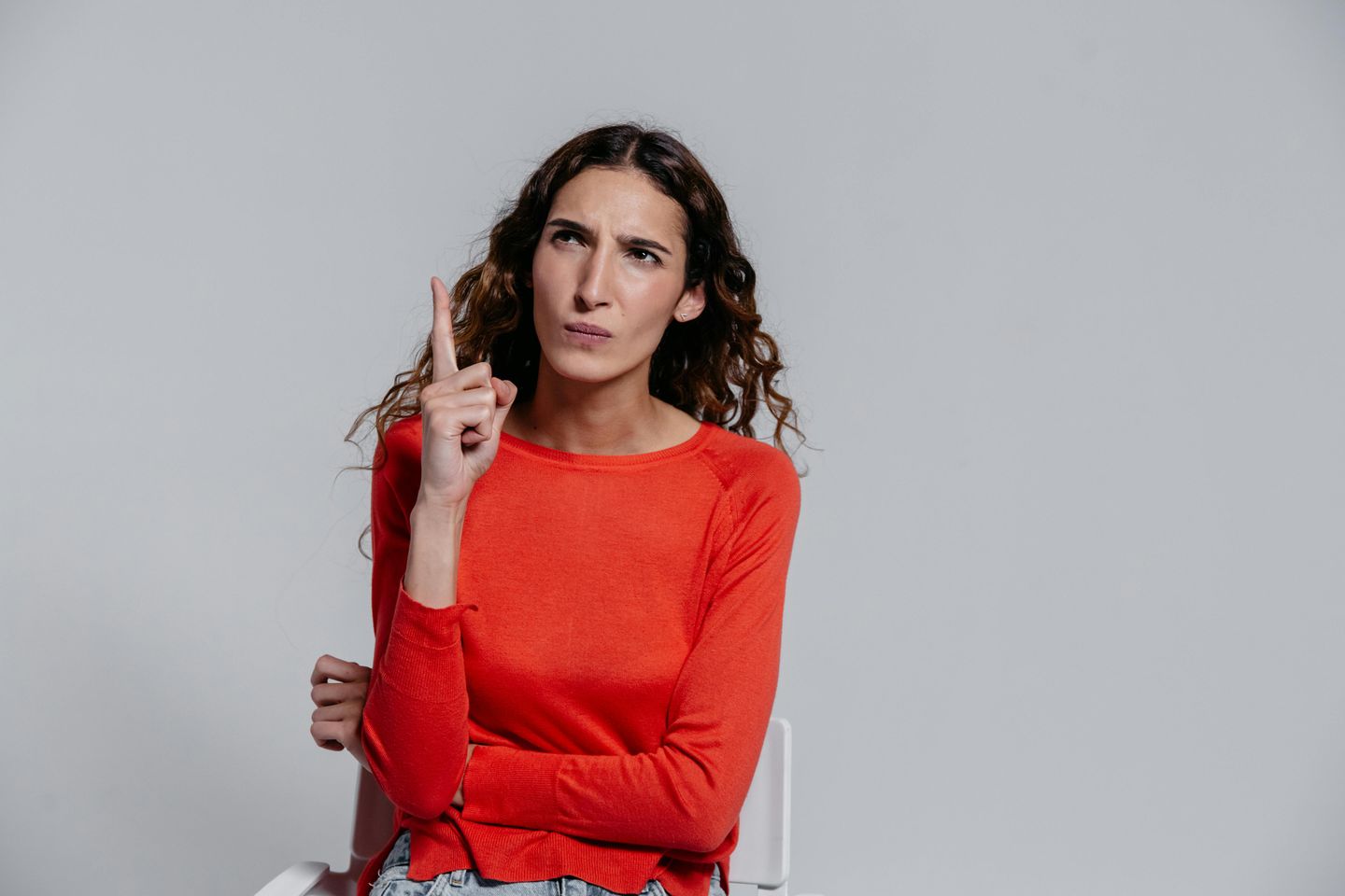 Mulher usando blusa vermelha faz expressão de dúvida enquanto levanta o dedo indicador, em fundo neutro.