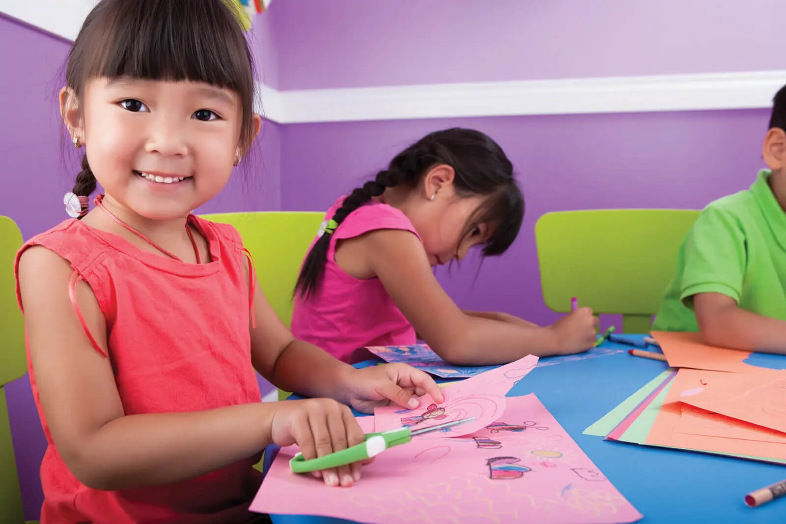 Children sitting at table cutting card while child in foreground smiles at camera