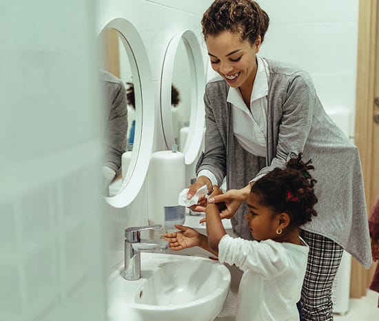 A woman helps a young girl wash her hands at a bathroom sink with a white oval mirror and a soap dispenser.