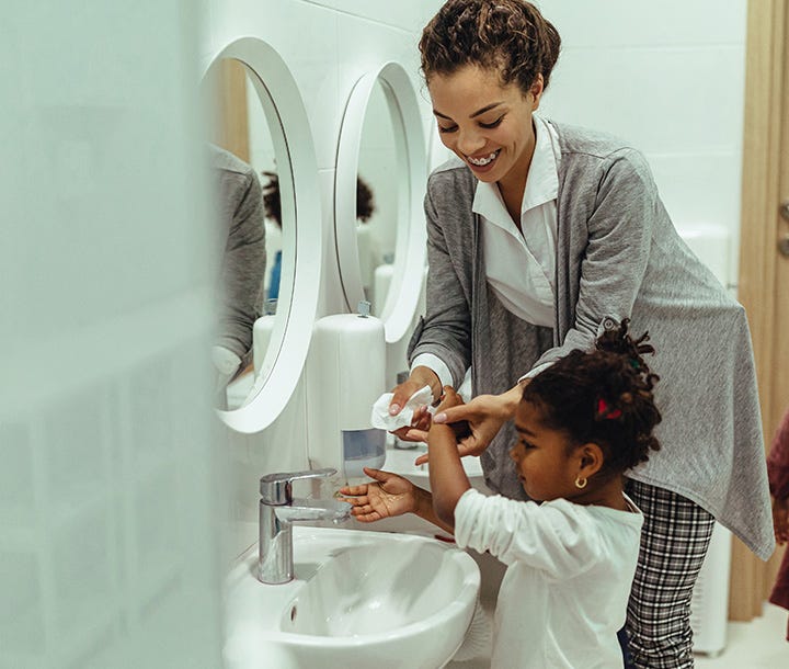 A woman helps a young girl wash her hands at a bathroom sink with a white oval mirror and a soap dispenser.
